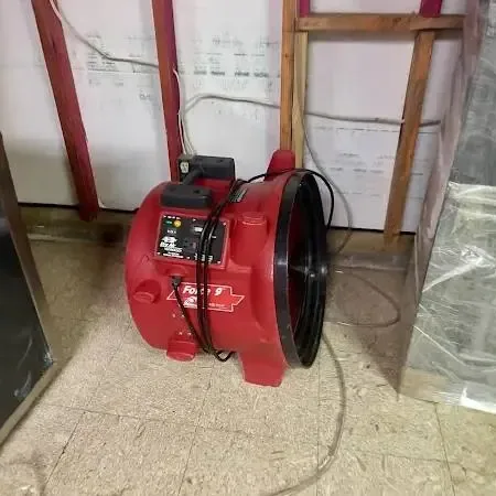 Red air mover running beside a room wall opened at the bottom, with plastic sheeting hung to contain the drying area
