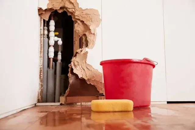 Wall opened around failed supply pipes with water on the floor and a bucket